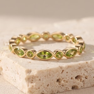 May include: A gold band ring featuring an alternating pattern of marquise-cut and round-cut light green peridot gemstones. The stones are set in a continuous row around the entire circumference of the metal band, creating a delicate and symmetrical design.