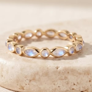May include: A gold ring featuring an alternating pattern of round and marquise-shaped moonstones set in a continuous band. The stones exhibit a soft blue iridescent glow, and the metal band has a polished finish, creating a delicate appearance.