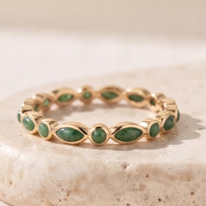 May include: A gold band ring featuring an alternating pattern of small round and marquise-shaped green stones set along the entire circumference. The metal has a polished finish, and the stones are evenly spaced to create a continuous design.