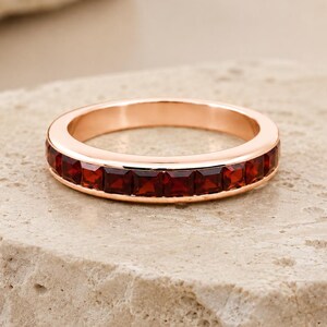 Puede incluir: Un anillo de banda en oro rosa con un engaste de canal de piedras preciosas granates de color rojo intenso y corte cuadrado que envuelven la circunferencia de la banda metálica, creando una línea continua de piedras carmesí en un marco de oro rosa pulido.