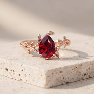 Könnte beinhalten: Ein Ring aus Roségold mit einem birnenförmigen, tiefroten Edelstein als Mittelstein. Die Ringschiene ist mit filigranen Metallblättern und Ranken gestaltet, die von der Fassung ausgehen und sich mit einer polierten Oberfläche um den Finger legen.