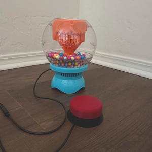May include: A bubble machine with a clear spherical container, an orange internal mechanism, and a light blue base. The machine is filled with small, multicolored spheres and is connected by a black cord to a round, red push-button switch.