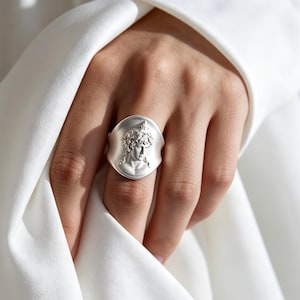 Peut inclure: Une bague argentée présentant un relief détaillé d'un buste classique aux cheveux bouclés. La bague possède un anneau large, lisse et légèrement concave qui encadre le portrait central, créant une apparence audacieuse et sculpturale.