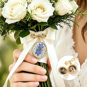 Può includere: Un ciondolo a medaglione circolare color oro fissato a un nastro bianco su un bouquet di rose bianche. Il medaglione presenta un motivo floreale blu sul davanti e si apre per rivelare due cornici circolari con ritratti di una coppia.