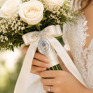 Peut inclure: Un bouquet de mariée composé de roses blanches et de gypsophile, noué par un ruban de satin crème. Un médaillon ovale en argent orné d'une petite fleur bleue est fixé au ruban par une épingle en forme de nœud argenté sur les tiges.