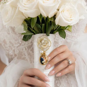 Op de afbeelding: Een boeket witte rozen met groene bladeren gewikkeld in een wit lint. Aan het lint is een goudkleurige metalen bedel bevestigd met een bloemmotief, een kleine witte parel en een ovaal hangertje met een foto van een koppel.