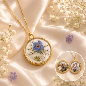 May include: A gold-toned circular locket necklace featuring a pressed blue and white flower arrangement on the front cover. The locket opens to reveal two circular photo frames containing portraits of a couple in wedding attire.