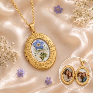 Könnte beinhalten: Ein ovales goldenes Medaillon mit geprägtem Blumenrand und einer gepressten blauen Blume im Inneren. Ein kreisförmiger Ausschnitt zeigt das geöffnete Medaillon mit zwei Innenrahmen, die jeweils ein Foto von zwei lächelnden Personen enthalten.