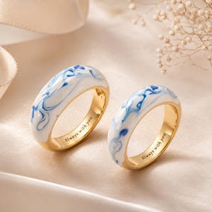 May include: Two gold-toned rings featuring a smooth, white enamel inlay with swirling blue patterns. The interior of each band is engraved with the text Always with you in a clean, serif font, showcasing a polished metallic finish throughout.
