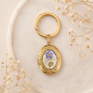 May include: A gold-toned oval locket keychain featuring an intricate embossed floral border. The center of the locket displays a white oval inlay decorated with a single pressed blue flower and a small white dried botanical element under a clear finish.