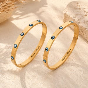 May include: Two gold-toned metal bangle bracelets featuring a series of small, circular blue and white evil eye charms embedded along the outer surface. Each bracelet is engraved with the black text Always with you on the inner band.
