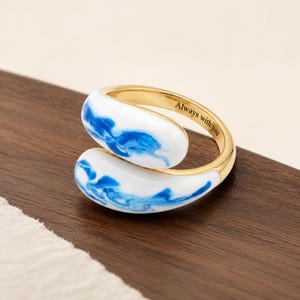 May include: A gold-toned open ring featuring two rounded, teardrop-shaped ends with a white and blue marbled enamel finish. The inner band of the ring is engraved with the black text Always with you, creating a distinct contrast against the metal.