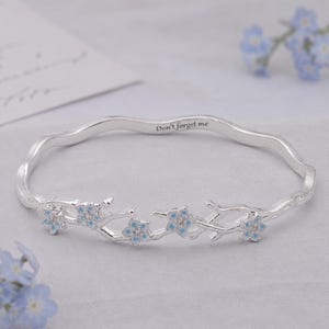 Könnte beinhalten: Ein silberner Armreif mit gewelltem Banddesign und einem Astmotiv, das mit kleinen hellblauen und weißen Blüten verziert ist. Die Innenseite des Reifs ist mit dem Text Don't forget me in einer klaren, dunklen serifenlosen Schrift graviert.