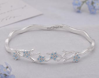 Op de afbeelding: Een zilveren armband met een golvend ontwerp en een takmotief versierd met kleine lichtblauwe en witte bloemen. De binnenkant van de band is gegraveerd met de tekst Don't forget me in een duidelijk, donker, schreefloos lettertype.