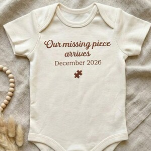 May include: Cream-colored baby onesie with short sleeves. The text "Our missing piece arrives December 2026" is printed in brown script, along with a brown puzzle piece graphic. A baby garment.