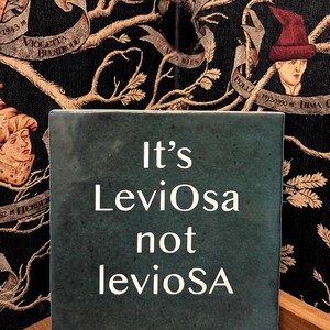 Puede incluir: Un azulejo de cerámica cuadrado de color verde oscuro con el texto blanco "It's LeviOsa not levioSA" centrado en la parte frontal. El azulejo tiene un acabado liso y ligeramente moteado, colocado sobre un fondo oscuro con motivos.