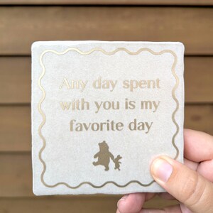 Könnte beinhalten: Ein quadratischer, heller Steinuntersetzer mit einem gewellten goldenen Rand. Der Text Any day spent with you is my favorite day ist in Gold gedruckt. Unter dem Text befindet sich eine kleine goldene Silhouette zweier Zeichentrickfiguren.