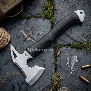 Pode incluir: Um machado tático com um cabo preto texturizado e uma lâmina prateada. A lâmina tem um formato único com uma borda afiada e uma seção dentada. O texto "Tjbikergear" é visível no machado. O machado está sobre uma superfície de pedra escura.