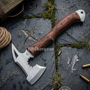 Pode incluir: Um machado tático com cabo de madeira e lâmina de aço inoxidável. A cabeça do machado apresenta uma lâmina afiada, uma ponta e uma seção dentada. O cabo é de cor castanho escuro. O texto "tjbikergear" é visível.