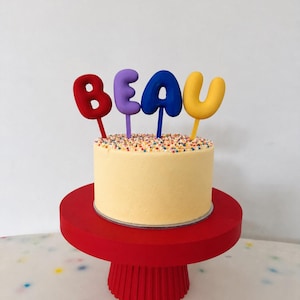 May include: A small, round, pale yellow cake topped with multi-coloured sprinkles and four individual letter-shaped toppers spelling the name BEAU in red, purple, blue, and yellow, all resting on a bright red, pedestal-style cake stand.