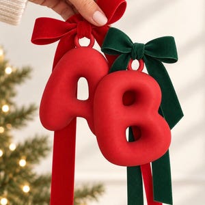 May include: Two red, matte-finish, three-dimensional letter ornaments, 'A' and 'B', each featuring a smooth, rounded surface. The letter 'A' is adorned with a red velvet ribbon bow, while the letter 'B' is adorned with a dark green velvet ribbon bow.