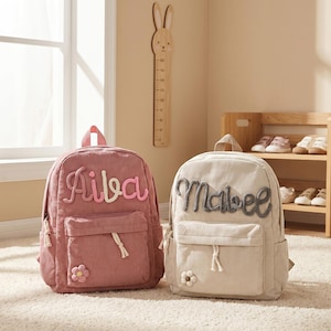 Könnte beinhalten: Zwei Rucksäcke aus Cord, einer in Staubrosa und einer in Creme, jeweils mit einem aufgestickten Namen auf der Vorderseite. Der staubrosa Rucksack hat den Namen "Aiba" in Rosa und Weiß, und der cremefarbene Rucksack hat den Namen "Mabel" in Grau. Beide haben eine Vordertasche und eine Blume.
