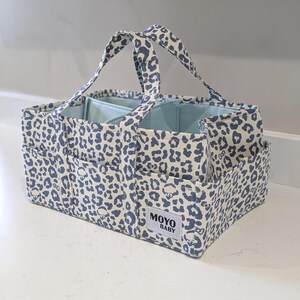 May include: A rectangular fabric storage caddy featuring a blue and white leopard print pattern. The caddy has two top handles, multiple exterior pockets, and an interior divider. A white rectangular label on the front displays the text MOYO BABY.