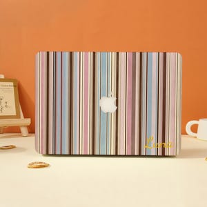 May include: A laptop case with a vertical striped pattern in pastel colors: pink, blue, brown, and white. The Apple logo is visible in the center. The name "Luna" is written in gold script on the lower right corner.