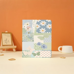 May include: A rectangular notebook featuring a patchwork design of light blue, green, and white panels. The pattern includes illustrations of white daisies, various green foliage, and several butterflies in shades of green and cream.