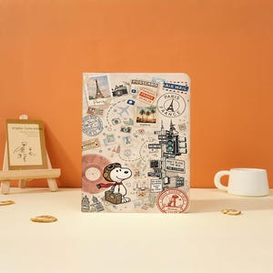May include: A rectangular notebook featuring a travel-themed design with a cartoon dog wearing goggles and a scarf, holding a suitcase. The cover displays illustrations of the Eiffel Tower, cameras, postage stamps, and street signs with text.