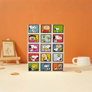 Puede incluir: Un cuaderno blanco con una cuadrícula de coloridos diseños de sellos postales de Snoopy. Cada sello muestra a Snoopy y otros personajes de Peanuts en varias poses. Una taza blanca y un pequeño caballete están en el fondo.