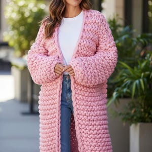 May include: A long, chunky knit cardigan in a soft pink colour. The open-front sweater features a thick, textured stitch and long sleeves. The cardigan is worn over a white top and blue jeans, creating a casual, comfortable look.