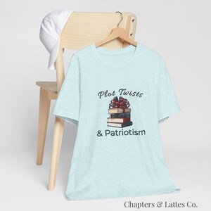 May include: Light blue t-shirt with the text "Plot Twists & Patriotism" and a graphic of a stack of books tied with a red bow. The shirt is hanging on a wooden hanger.
