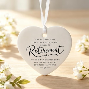 May include: A white heart-shaped ceramic ornament hanging from a thin white ribbon. The text reads: SAY GOODBYE TO THE ALARM CLOCKS AND HELLO TO RETIREMENT. MAY THIS NEW CHAPTER BRING YOU JOY, FREEDOM AND COUNTLESS HAPPY DAYS, with small heart icons.