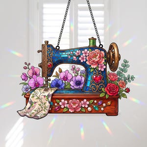 Könnte beinhalten: Ein dekoratives Hängeornament in Form einer Vintage-Nähmaschine mit einem blauen Körper im Buntglas-Stil, verziert mit rosa Pfingstrosen, roten Rosen und violetten Orchideen. Ein Stoffstück mit Kolibri-Illustrationen hängt an der Nadel.