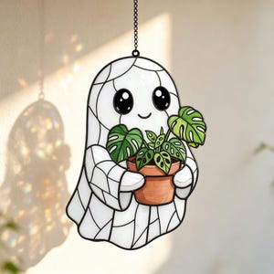 May include: A stained glass suncatcher shaped like a white ghost with black outlines, large black eyes, and a small smile. The ghost holds a terracotta-coloured pot containing a green monstera plant with detailed leaf cutouts and black lead lines.