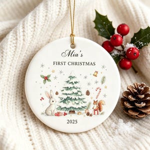 May include: A round white ceramic ornament featuring a gold hanging string. The design includes a central snow-covered Christmas tree, a rabbit, a squirrel, gifts, and the text "Mia's FIRST CHRISTMAS" at the top and "2025" at the bottom.