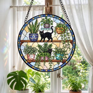 Op de afbeelding: Een ronde glas-in-lood zonnevanger met twee zwarte katten tussen diverse potplanten op houten planken. Het ontwerp bevat een sansevieria, een hangende vetplant en ander groen in kleurrijke potten, omlijst door een blauwe en gele rand.