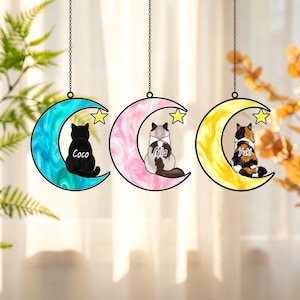 May include: Three hanging crescent moon ornaments with black metal chains. Each moon features a marbled color—blue, pink, and yellow—and a small yellow star. A cat sits on each moon with the names Coco, Mia, and Yuki printed on their backs.