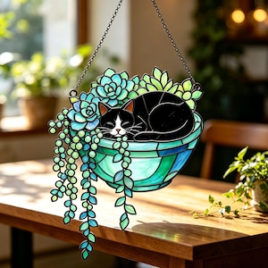 Op de afbeelding: Een hangende glas-in-lood zonnevanger met een zwart-witte kat die opgerold in een blauwgroene kom ligt, gevuld met groene vetplanten en hangende ranken. Het object hangt aan een dunne zwarte metalen ketting met donkere loden omlijningen.