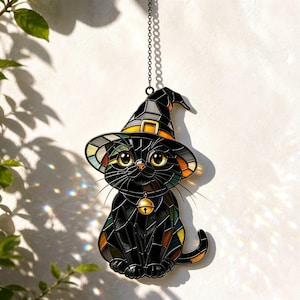 Op de afbeelding: Een glas-in-lood zonnevanger in de vorm van een zwarte kat met een puntige heksenhoed. De kat heeft grote gele ogen en een halsband met een gouden belletje. De hoed is voorzien van een gele band en oranje accenten.