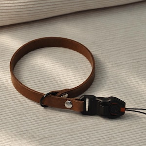 Könnte beinhalten: Ein braunes Leder-Handgelenkband mit kreisförmigem Design. Das Band ist mit zwei silberfarbenen Metallnieten befestigt und verfügt über einen schwarzen Kunststoff-Schnellverschluss mit einer angebrachten dünnen schwarzen Kordelschlaufe.