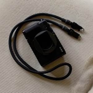 Pode incluir: Uma câmara digital preta com um para-sol quadrado e o texto X-E4 impresso no corpo frontal. A câmara está ligada a uma correia de pescoço comprida, redonda e preta, estilo corda, com dois conectores metálicos pretos nas extremidades.