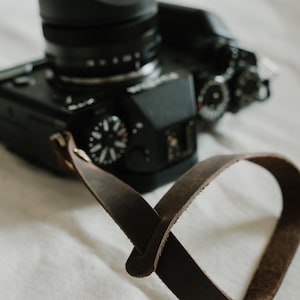 May include: A dark brown leather camera strap with a smooth finish and a simple loop attachment mechanism, connected to a black digital camera body featuring various manual control dials and a lens with a black hood attached to the front.