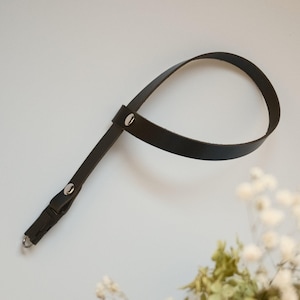 Op de afbeelding: Een zwarte leren riem gevormd tot een lus, vastgezet met twee zilverkleurige metalen klinknagels. De riem heeft een kort uiteinde met een kleine zilverkleurige metalen ring, wat een functioneel bevestigingspunt biedt voor diverse voorwerpen.