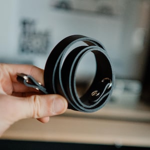 Könnte beinhalten: Ein aufgerolltes schwarzes Lederarmband mit silberfarbener Hardware. Das Armband wird in einer Hand gehalten, wodurch seine glatte Textur und sein Design zur Geltung kommen. Der Hintergrund ist unscharf, wodurch die Aufmerksamkeit auf das Armband gelenkt wird.