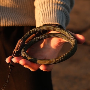 May include: A coiled olive green rope wrist strap with a brown leather accent and a black clasp. The rope has a textured, braided appearance. The strap is held in a hand, with a neutral background.