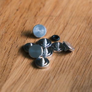May include: A collection of small, circular silver-colored metal studs with pointed threaded backs, featuring a smooth flat top surface. One tiny black rubber O-ring is included among the metallic hardware components, which are arranged in a cluster.