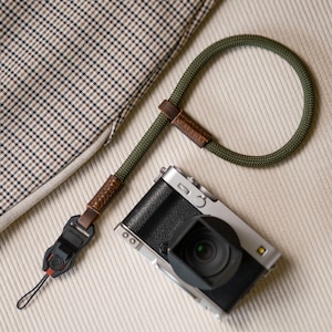 Può includere: Una tracolla per fotocamera in corda intrecciata verde oliva con dettagli in pelle marrone e un connettore rapido in plastica nera, fissata a una fotocamera digitale argento e nera con paraluce, accanto a un tessuto a quadri beige.