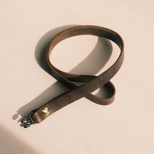 Pode incluir: Uma pulseira de couro castanho com fecho de metal e um rebite dourado. A pulseira está em laço e repousa sobre uma superfície de cor clara. O couro tem um aspeto texturizado.